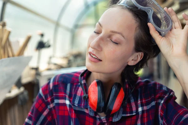 Young beautiful handy professional happy female strong carpenter portrait wearing protective goggles working in carpentry diy workshop against wood. Confident engineer. Women male hobby at workbench