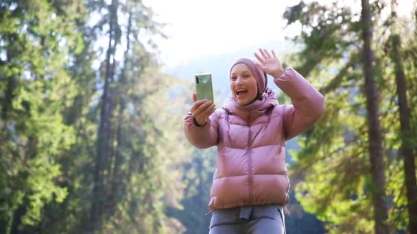 Νεαρή ενήλικη καυκάσια όμορφη γυναίκα σε ζεστό σακάκι και μαλλί κοστούμι βίντεο chating με κινητό τηλέφωνο σε φίλους ή την οικογένεια κατά τη διάρκεια του περπατήματος σε γραφικό δάσος πεύκο δάσος.Πρόσωπο κάνει selfie περπάτημα σε εξωτερικούς χώρους — Αρχείο Βίντεο