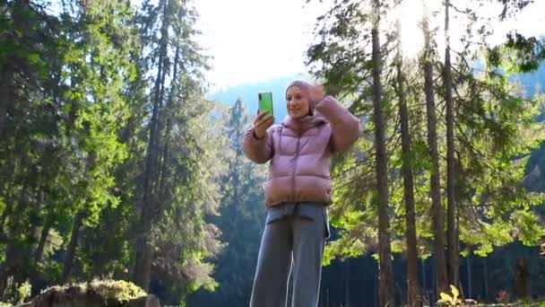 Mladý dospělý kavkazský krásný žena v teplé bundy a vlna oblek video chatování mobilním telefonem přátelům nebo rodině během procházky v malebném zeleném lese borovic.Osoba, aby selfie chůze venku — Stock video