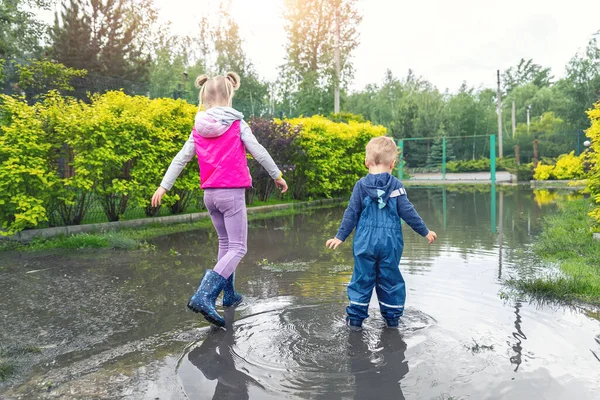 Bakåt bild av två söta blonda kaukasiska små barn bror och syster njuta av att spela hoppa i smutsiga pöl bär blå vattentäta byxor och gummi regnstövlar på gatan utomhus — Stockfoto