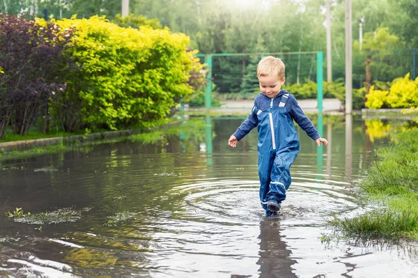 Pequeño niño pequeño rubio caucásico juguetón disfrutar de divertirse jugando saltar en charco sucio con pantalones azules impermeables y bota de lluvia de goma en casa patio calle al aire libre. Concepto de infancia feliz — Foto de Stock