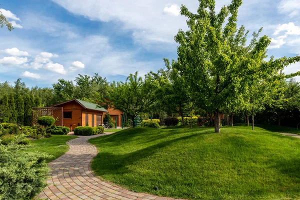 Vista panoramica giardino alberi da giardino e strada asfaltata sentiero in pietra per camminare contro il bel cielo blu. Progettazione del paesaggio verde prato erboso colline e piante irrigate con spruzzatori autonomi intelligenti nella giornata luminosa — Foto Stock