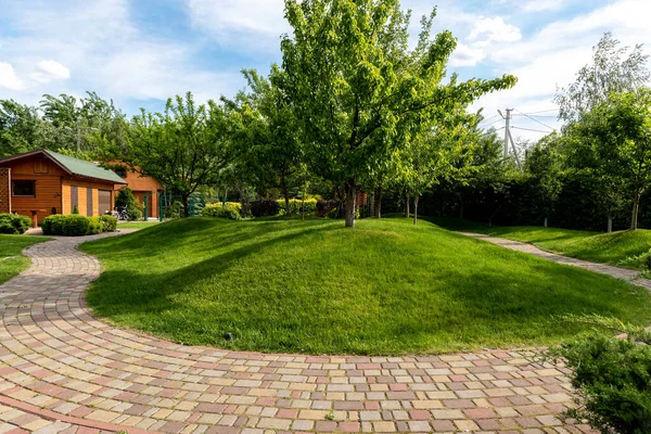 Vistas panorámicas jardín jardín árboles y camino de piedra pavimentada para caminar contra el hermoso cielo azul. Diseño de paisaje verde césped césped colinas y plantas regadas con pulverizadores autónomos inteligentes en el día brillante — Foto de Stock