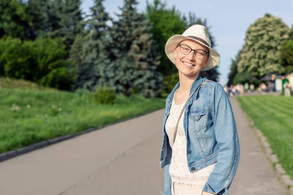 Feliz joven mujer calva caucásica con sombrero y ropa casual disfrutando de la vida después de sobrevivir al cáncer de mama. Retrato de la hermosa chica sin pelo sonriendo durante el paseo en el parque de la ciudad después de curar la enfermedad —  Fotos de Stock