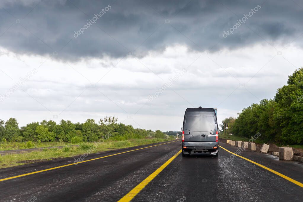 Back of delivery small shipment cargo courier van moving fast on wet highway road to city urban suburb against rainy cloudy sky . Business distribution and logistics express service. Mini bus motion