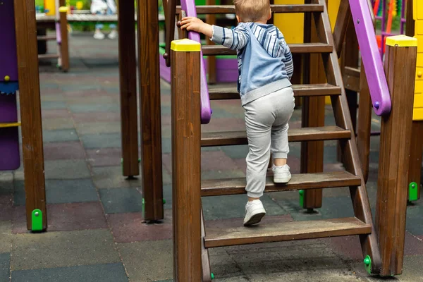 Vista posterior lindo adorable caucásico rubio niño pequeño niño divirtiéndose disfrutar de subir escaleras de madera en los niños moderno parque infantil en el día de verano brillante. Al aire libre niños calle deporte actividades —  Fotos de Stock