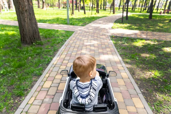 Aranyos kis kaukázusi szőke kisgyerek élvezni szórakozás lovaglás elektromos meghajtású játék autó aszfalt út út városi park nyári napon. Boldog gyermek közel útkereszteződés döntéshozatal jövőbeli koncepció — Stock Fotó