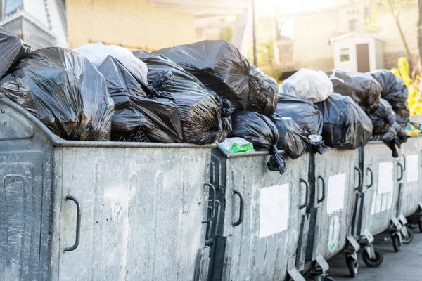 Filas de muchas latas de basura de metal sobrecargado llenas de bolsas de basura de plástico negro cerca de un edificio residencial en el centro de la ciudad o área suburbana. Recogida de basura de clasificación no reciclable — Foto de Stock