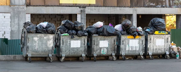 Linhas de muitas latas de lixeira sobrecarregadas de metal grande cheias de sacos de lixo de plástico preto perto de edifício residencial no centro da cidade ou área suburbana. Coleta de lixo de triagem não reciclável — Fotografia de Stock