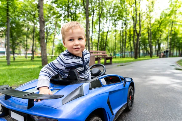 Portré aranyos kis kaukázusi szőke kisgyerek élvezi a szórakozást lovaglás elektromos meghajtású játék autó aszfalt út út városi park nyári napon. Boldog gyermek élvezze játszani rc jármű a szabadban — Stock Fotó