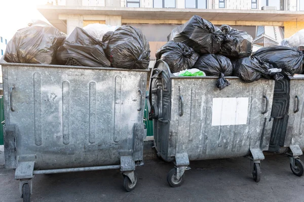 Filas de muchas latas de basura de metal sobrecargado llenas de bolsas de basura de plástico negro cerca de un edificio residencial en el centro de la ciudad o área suburbana. Recogida de basura de clasificación no reciclable — Foto de Stock