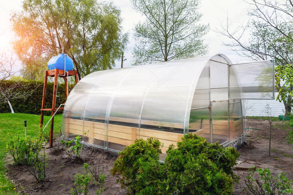 DIY wooden base with elevated water storage tank for autonomous drip irrigation of garden bed and growing green fresh organic vegetable in eco-friendly small greenhouse yard. Domestic farming concept