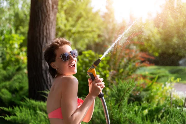 Fiatal felnőtt szexi karcsú sportos kaukázusi barna nő élvezni szórakozás locsoló kert otthon udvar tömlő cső zuhany forró nyári napon. Boldog női személy pihenjen a kertben park szabadban — Stock Fotó