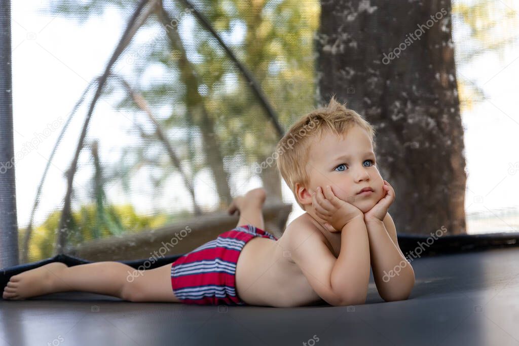 Portrait of cute bored little caucasian funny sad blond toddler boy lying inside big black trampoline at home backyard playground area outdoors warm summer sunny day. Children street sport activity