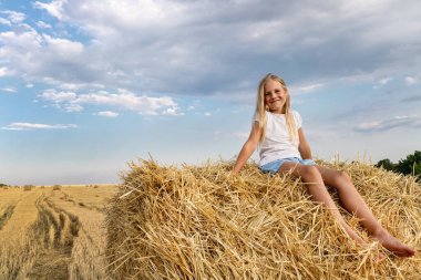 Şirin, küçük sarışın, sevimli, sevimli beyaz kız çocuğunun portresi samanlıkta oturmaktan ya da hasat edilen buğday tarlasında sıcak bir yaz akşamında balyada oturmaktan zevk alır. Kırsal arazi manzarası