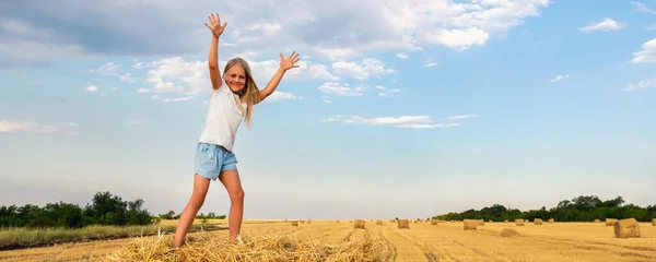 Porträtt av söt liten blond vacker bedårande glad kaukasiska unge flicka njuta av att sitta på höstack eller balar på skördade vetefält varm sommarkväll. Natursköna landsbygd landskap bakgrund — Stockfoto