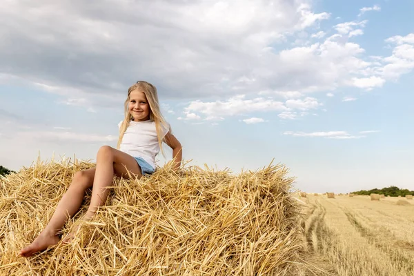 Portrait of cute little blond beautiful adorable cheerful caucasian kid girl enjoy sitting on hay stack or bale on harvested wheat field warm summer evening. Scenic rural country landscape background — Stock Photo, Image