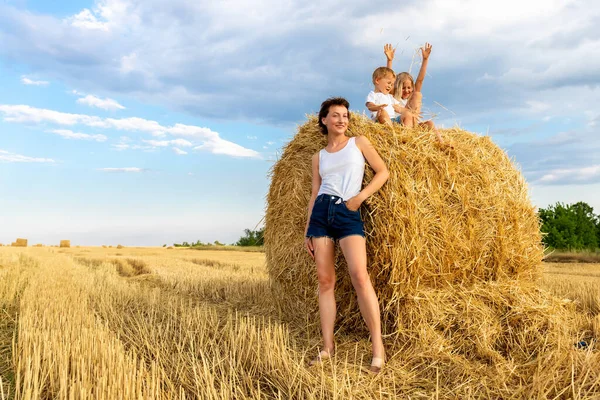 Mladý dospělý atraktivní krásná maminka s malým synem a dcerou se baví flákání kolem sedí u zlatého sena balík na pšeničné sklizené pole v blízkosti farmy. Šťastné děti na venkově — Stock fotografie