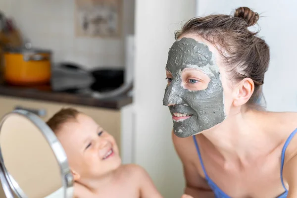 Young adult happy beautiful caucasian woman with little son applying natural gray clay face mask at home against kitchen hob at day time. Female person making self facial cleansing moisturizing care — Stock Photo, Image