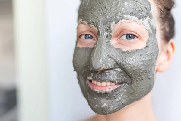 Joven adulto feliz hermosa mujer caucásica aplicando mascarilla de arcilla gris natural en casa contra la ventana y el espejo en el día. Persona femenina haciendo auto limpieza facial hidratante tratamiento de cuidado —  Fotos de Stock