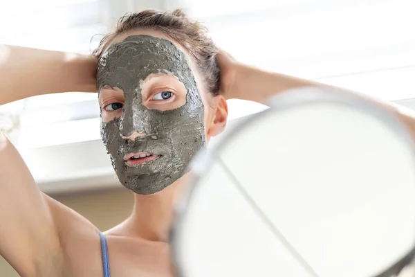 Joven adulto feliz hermosa mujer caucásica aplicando mascarilla de arcilla gris natural en casa contra la ventana y el espejo en el día. Persona femenina haciendo auto limpieza facial hidratante tratamiento de cuidado —  Fotos de Stock