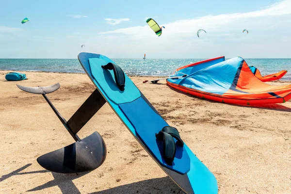 Close-up hydrofoil surf board and kite equipment on sand beach shore watersport spot on bright sunny day against sea ocean coast with many kiter riding surf school camp. Active travel sport concept.