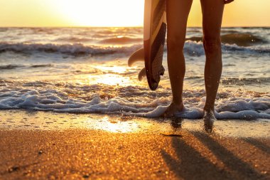 Dibe yakın çekim genç yetişkin sörfçü kız bacakları okyanus kıyısında güneş doğarken ya da günbatımında sörf tahtası üzerinde duruyor. Spor sağlıklı kaygısız özgürlük yaşam tarzı tatil konsepti