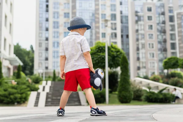 Visão traseira de bonito sozinho triste adorável caucasiano 3-4 anos loiro menino em shorts elegantes camisa e chapéu segurar brinquedo de pelúcia macio perdido ao ar livre no centro da cidade urbana da área. Criança solitária perturbada — Fotografia de Stock