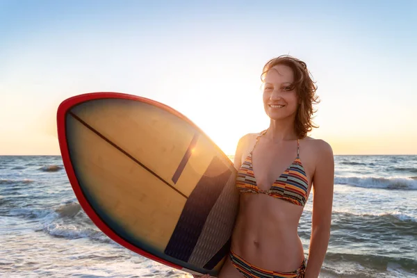 Portret jong volwassen slanke sportieve vrouwelijke surfer meisje met surfplank silhouet staan aan de oceaankust golf tegen warme zonsopkomst of zonsondergang zon. Sport gezonde zorgeloze vrijheid levensstijl vakantie concept — Stockfoto