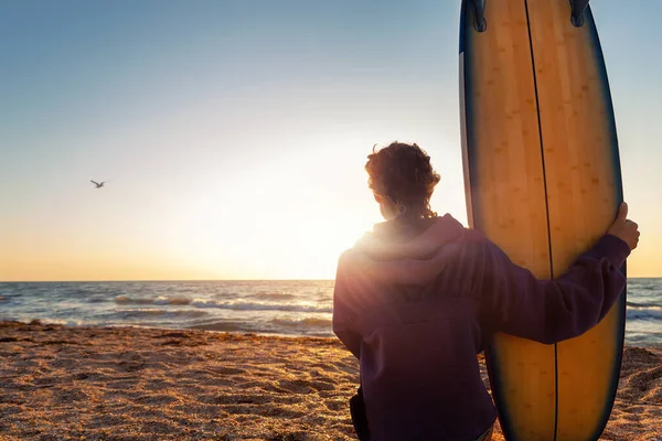 Retrovisore giovane adulto sottile ragazza surfista sportiva con tavola da surf seduto sulla sabbia a onda costa oceanica contro l'alba calda o il sole del tramonto. Sport sano spensierato libertà stile di vita vacanza concetto — Foto Stock