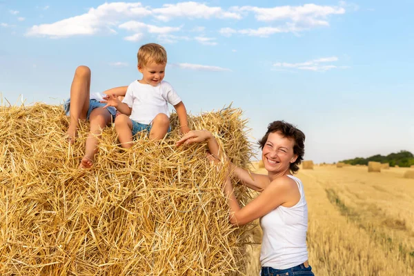 Ung vuxen attraktiv vacker mamma med liten son och dotter njuta av att ha kul lura runt sitter nära gyllene höbalar på vete skördade fält nära gården. Glada barn på landsbygden — Stockfoto