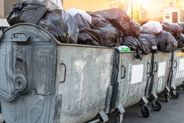 Filas de muchas latas de basura de metal sobrecargado llenas de bolsas de basura de plástico negro cerca de un edificio residencial en el centro de la ciudad o área suburbana. Recogida de basura de clasificación no reciclable — Foto de Stock