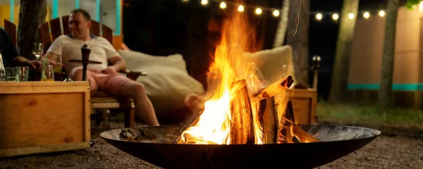 Fuoco in ghisa posto falò presso campeggio spiaggia foresta con brgiht fiamma ardente di sera contro ghirlanda lampadina e alberi. Amici sedersi vicino falò campo stufa a calda notte d'estate — Foto Stock