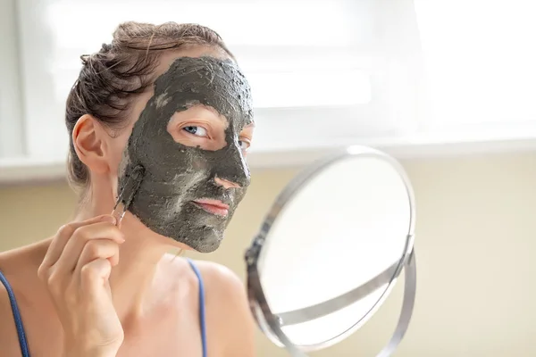 Joven adulto feliz hermosa mujer caucásica aplicando mascarilla de arcilla gris natural en casa contra la ventana y el espejo en el día. Persona femenina haciendo auto limpieza facial hidratante tratamiento de cuidado —  Fotos de Stock