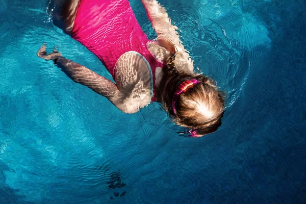 Felül a felső nézet a kis aranyos gyerek lány sport rózsaszín fürdőruha és szemüveg búvárkodás víz alatt tiszta kék víz fedett medence sötét este. Gyermekek vízi sportok egészséges tevékenységek — Stock Fotó
