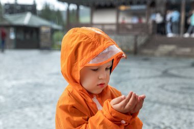 Portre, parlak turuncu su geçirmez yağmurluk giyen beyaz çocuk yağmurluk giyer ve dışarıdaki Avrupa caddesinde ellere düşüşün keyfini çıkarır. Sonbahar havası açık havada yürüyor.
