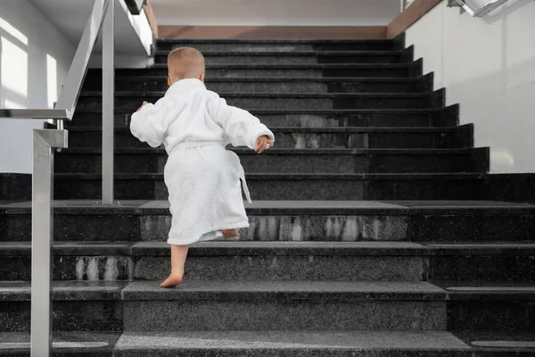 Back view of cute funny caucasian blond kid boy wearing white pool spa bathrobe walking upstairs on big marble staircase. Alone small child meet challenge and purpose. Rich luxury lifestyle concept — Stock Photo, Image