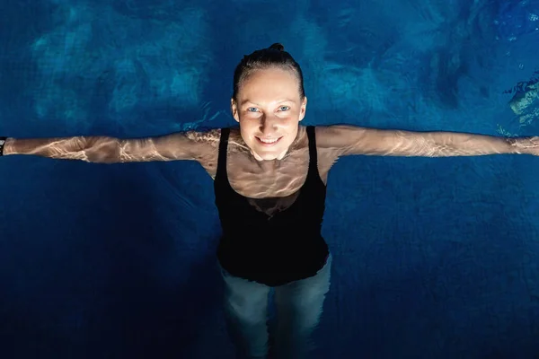 Портрет молодої дорослої кавказької моделі в спортивному чорному купальнику стоїть у чистій блакитній воді критого басейну в темний вечірній час доби. Розслабте гармонію та оздоровчу спа-терапію — стокове фото