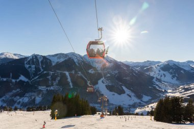 Alp yamacında güneşli bir kış günü teleferik istasyonunda kayak ve snowboard sporları dinleniyor. Parlak güneş ışığı, mavi gökyüzü ve dağ manzarası macera, özgürlük ve rahatlama atmosferi yaratır..