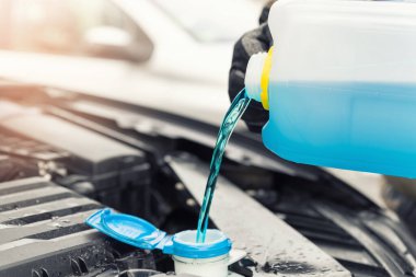 Close-up detail of driver or mechanic hand in gloves pouring blue antifreeze liquid for winter car windshield screen washing with bootle watering can. Car seasonal service and maintenance concept.
