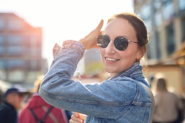Portre genç yetişkin oldukça mutlu kadın şehir sokaklarında yürüyüş yaparken eğleniyor güneşli bir akşam. Denim ceket çiçekli güneş gözlüğü neşeli enerjiyle dolu canlı bir atmosfer yaratır..