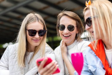 Gülümseyen arkadaşlar birlikte telefona bakar, neşeli ifadeler ve rahatlamış ruh halleriyle paylaşılan anın tadını çıkarırlar. Günlük tarz, güneş gözlüğü ve canlı renkler, neşeli bir bağlantı ortamı yaratır. Sosyal eğlence..