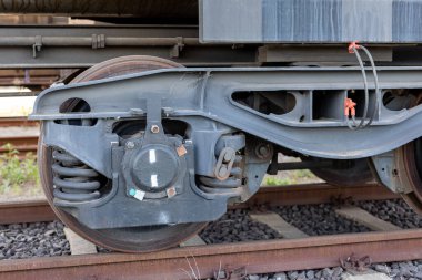 Ağır çelik yayları ve yıpranmış tekerlek yüzeyi olan metal yük vagonu gün ışığında paslı raylarda duruyor. Endüstriyel yapı süspansiyon, yön bulma ve görünür dokularla bağlantıları gösteriyor.