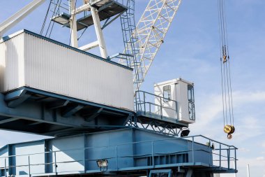 Endüstriyel vinç yapısı temiz gökyüzü altında yükseliyor, metal iskelet, kulübe, makara ve kanca sergiliyor. Sahne, makineleri, çelik geometriyi ve güçlü güneş ışığı ve gölgeli port atmosferini vurgular.