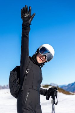 Portre genç, mutlu, gülümseyen kadın kayakçı siyah kıyafetler giyiyor. Kar yamacında duruyor. El miğferi takıyor. Soğuk, güneşli kış günü. Alp tatil köyü dağ manzarası Mavi gökyüzü.