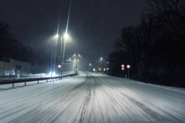 Şiddetli kar yağışı sırasında gece yolu karla kaplı sokak lambaları ve farlarla aydınlatılmış hareket bulanıklığı yaratıyor. Buzlu asfalt ve trafik işaretleri tehlikeli durumu vurguluyor.