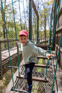 Gülümseyen çocuk, orman zemininin yukarısındaki halat köprüsü macera parkından geçiyor. Ahşap yapı etkinlik dengesi cesaret aile eğlencesi bahar doğası çocukluk eğlencesi.