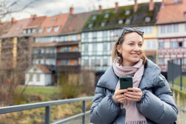 Avrupa 'nın tarihi şehrinde elinde akıllı telefon tutan gülümseyen kadın sıcak ceket ve eşarp giyerek sakin şehir havasının tadını çıkarıyor. Resim modern iletişimi, seyahat yaşam tarzını ve bağlantıyı temsil ediyor.