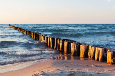 Ahşap dalgalar, yumuşak akşam ışığı altında mavi denize doğru uzanıyor. Yumuşak dalgalar kumlu sahile dokunurken, güneş sıcacık ahşap direklere ve parıldayan suya yansıyor..