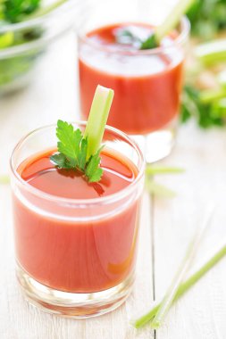 Two glasses with tomato juice, celery and parsley stand on a wooden table in white. Stalks of celery sliced on a cutting board in the background.
